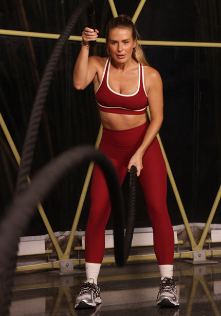 A woman works out with heavy battle ropes in a dark gym, wearing a red Lorna Jane sports bra and matching leggings with white sneakers.