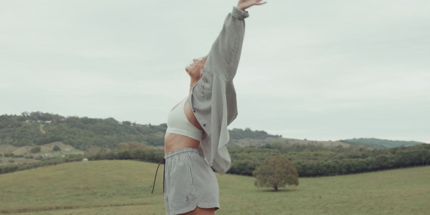 A woman moves through a rolling green valley and lush tropical garden in three elevated warm weather looks, a white crop top with gingham shorts and an oversized striped shirt, a white silk slip dress, and a textured white knit top with striped shorts. Lorna Jane lifestyle activewear.